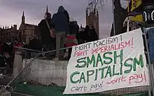 An einem umgeworfenen Bauzaun ist ein weißer Banner befestigt, auf dem die Sätze „Fight Racism!“ (in schwarzer Farbe), „Fight Imperialism!“ (in roter Farbe) sowie in Großbuchstaben „Smash Capitalism“ (in grün) steht. In etwas kleineren Buchstaben steht noch „Can't pay! Won't Pay“ (erster Satz in schwarz, zweiter in rot) am unteren Rand des Banners. Links hinter dem Banner steht eine Gruppe junger Menschen.
