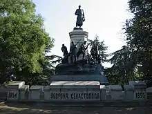 Frontale Farbfotografie eines Denkmals mit kyrillischem Titel und den Jahreszahlen 1854 und 1855 auf einer Steinmauer. Der untere Sockel hat einen gegossenen Felsen mit einigen Soldatenfiguren darauf, die einen weiteren Sockel mit einem General umkreisen. An beiden Seiten des Denkmals sind Bäume.