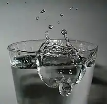Kurzzeitfotografie eines mit Wasser gefüllten Trinkglases. Die Wasseroberfläche ist aufgewirbelt, ein Loch von etwa 3 cm Durchmesser ist im Wasser. Am Rand des Loches befindet sich ein Kraterrand, der sich etwa 2 cm über die restliche Wasseroberfläche erhebt. Einzelne Wassertropfen haben sich vom Kraterrand gelöst und befinden sich in etwa 4 cm über dem Rand des Glases. Vom Kraterrand laufen zwei dünne Arme aus Wasser nach oben, in einzelne Tröpfchen zu zerfallen.