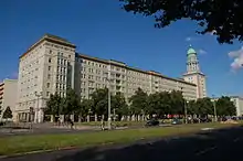 Seitliche Farbfotografie eines mehrstöckigen Gebäudes mit einem Turm an der rechten Seite. Der Turm hat einen runden Säulenaufbau und eine grüne Kuppel. Das Gebäude hat einen verzierten Dachgesims und vier kleine Vorbauten. Im Erdgeschoss sind Geschäfte, die von Bäumen verdeckt sind.