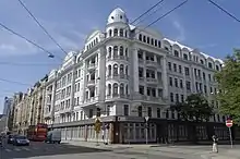 Eckansicht in Farbe von einem weißen, mehrgeschossigen Gebäude mit runder Ecke und großen Balkonen. Als Verzierung dienen Säulen und unter den Dachsimsen sind Ornamente. Über den obersten Fenstern sind Giebelfelder und auf der Spitze der Eckfenster ist eine blaue Kuppel.