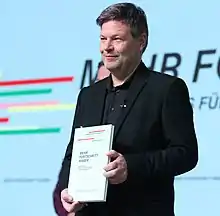 Seitliche Farbfotografie von Habeck, der auf einer Bühne lächelnd ein Buch mit dem Titel „Mehr Fortschritt wagen“  im DIN-A4-Format vor seinem Körper hält. Er trägt einen schwarzen Anzug und ein schwarzes Hemd. Im Hintergrund ist das Buchcover auf der großen Leinwand.