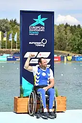 Roman Poljanskyj im Rollstuhl vor der Regattastrecke Oberschleißheim. Er trägt blaue Sportkleidung über einem weißen T-Shirt. Die rechte Faust reckt er geballt in die Höhe.
