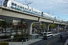 Verkehrswege in der Stadt mit viel Asphalt und Beton, Bürogebäude im Hintergrund. Obere Bildhälfte Stahlbetonviadukt, darauf eine Vierfachtraktion aus Doppeltriebwagen im Sound-Transit-Farbschema weiß-hellblau mit dunklerem meereswellenartigen Farbverlauf. Darunter an Bushaltestelle mit größerem verglasten Unterstand ein aufgrund der Perspektive klein wirkender Doppelstockbus ebenfalls im Sound-Transit-Farbschema.