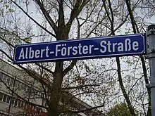 Blaues Straßenschild mit weißer Schrift. Aufschrift: Albert-Förster-Straße. Dahinter sind Bäume zu sehen. In der linken unteren Hälfte ist der obere Teil von zwei Gebäuden abgebildet.