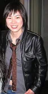 Alice Wu steht vor einer grauen Wand mit einem dunkelgrünen Vorhang. Sie trägt eine schwarze Lederjacke, ein graues Hemd, eine Jeans mit braunem Gürtel und eine dunkelbraune Krawatte mit weißen Punkten. Ihre schwarzen Haare trägt sie als strähnige, bis zum Hals reichende Bobfrisur mit einem fransigen Pony.