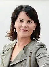 Frontalaufnahme von Baerbock, die direkt in die Kamera blickt. Dabei ist ihr linker Oberkörper nach vorne gerichtet und der linke Arm verschwindet unter der Kamera. Sie trägt einen olivgrünen Blazer und ihre dunkelbraun gefärbten Haare sind schulterlang.