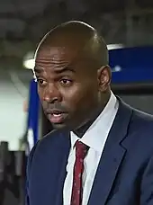 Nahaufnahme des in einem dunkelblauen Anzug, dunkelrotem dezent gemustertem Schlips und weißem Hemd mit geschlossenem Kragen gekleideten Antonio Delgado während einer Rede in einem Busdepot. Delgado steht leicht vorgebeugt hinter einem hier nicht sichtbaren Rednerpult und hat den Mund zum Sprechen geöffnet.
