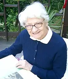 Asuman Baytop. Eine ältere Frau in Strickjacke im Stuhl mit einem Pflanzenbuch auf dem Schoß. 2003