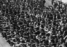 Schwarz-Weiß-Bild einer Menschenmenge (ca. 20 dichte Reihen, hauptsächlich Männer), in die linke untere Bildecke orientiert den Hitlergruß zeigend. Mitten im oberen rechten Quadranten des Bildes steht ein einzelner Mann mit verschränkten Armen, auf dem Foto durch einen grauen Kreis hervorgehoben.