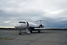 Privatjet des Herstellers Bombardier, Modell Global 5000, Seitenansicht, die Tür ist offen, die Treppe ausgeklappt, es sind elf Fenster auf der rechten Flugzeugseite zu sehen, das Flugzeug steht auf dem Vorfeld eines Flughafens, der Himmel ist trüb