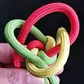 Mit dem nächsten Kardeel (gelb) kreuzt man über dem zuerst bewegten Kardeel (grün) und dem nächsten (rot). Dann noch über das zuerst bewegte Kardeel (grün) nach unten durch.