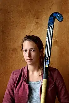 Nelen in grauem T-Shirt und weinroter Kostümjacke. Von unterhalb des Bildes bis weit über Nelens Kopf ist ein Hockeyschläger im Bild.