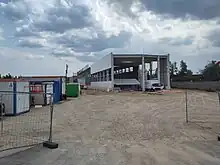 Die Betriebshalle ist im Rohbau zu sehen, einige Wände fehlen noch. Am linken Bildrand Baucontainer.