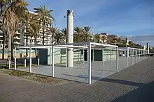 Foto einer Strandpromenade mit Palmen und Hotels, im Vordergrund zwei niedrige graugrüne Gebäude mit der Aufschrift Beach Club Six.