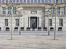 Frontale Farbfotografie einer Eingangstür aus Glas mit Säulen an beiden Seiten und einer ausgestreckt sitzenden Figur im oberen Relief. Die Figur und die Tür befinden sich in einer rechteckigen Nische, die von vielen Fenstern umgeben ist. Vor den unteren Fenstern befinden sich Gitterstäbe. Der Vorplatz ist mit Pflanzkübeln und vier kahlen Bäumen gesäumt.