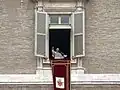 Farbfotografie von Papst Benedikt 16, der an einem durchsichtigen Rednerpult am offenen Fenster steht und seine rechte Hand hebt. Am Fenster sind graue Fensterläden und ein dunkelroter langer Stoff mit goldenen Verzierungen und das Wappen hängt am Fenstersims.