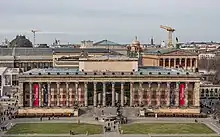 Frontale Farbfotografie eines dunklen Gebäudes mit einer durchgehenden Säulenvorhalle, hinter der sich ein breiter Glaseingang und rote Kachelwände befinden. Ein lateinischer Text steht im Gesims. Ein rechteckiger Aufbau mit Pferden- und Menschenfiguren sowie einer Fahne befindet sich auf der Dachmitte. Im Vordergrund ist eine Wiese mit einem Springbrunnen.