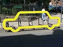 Eine hellgelbe Konstruktion in der Form eines stilisierten Autos, etwa anderthalb Meter hoch und fünf bis sechs Meter breit. Dahinter erkennt man mehrere, parallel aufgestellte Fahrradbügel. Die Konstruktion steht im Rand einer Straße, die Fahrradparkplätze sind durch das gelbe Auto vom Gehweg aus zugänglich. Dahinter sieht man einen kleinen Park und eine städtische Straße.