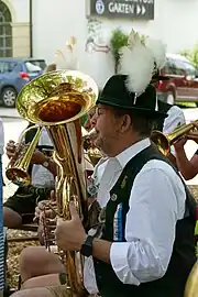 Foto des Oberkörpers eines Mannes, der in ein Blechblasinstrumen bläst. Er trägt einen grünen Hut mit Federn. Im Hintergrund sind weitere Musiker zu sehen