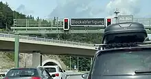 Eine Schilderbrücke quer über einer Straße. In der Mitte der Brücke ist eine leuchtende Anzeige "Blockabfertigung" angebracht, rechts und links davon je eine rot zeigende Ampel. Unter der Brücke stauen sich einige Autos. Im Hintergrund ist ein Tunnelportal zu erkennen.