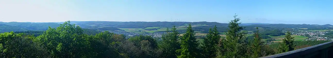 Die Hochflächen vom Koppeturm im Ostteil der Zollbuche aus etwa 470 m Höhe gesehen; links der Mitte die Angelburg (609 m; Sendemast), rechts der Mitte Daubhaus (552 m) und Allberg (528 m) am Ostrand, dazwischen ist das Relief auffällig unbewegt. Ganz rechts im Bild der Rimberg (497 m; Damshäuser Kuppen), links davon in hinterer Reihe das Arennest (592 m; Sackpfeifen-Vorhöhen) und die Sackpfeife (674 m); der Schwarzenberg (561 m; Breidenbacher Grund) wird bereits teilweise durch den Allberg verdeckt. Im zentralen Vordergrund Weidenhausen, im rechten Gladenbach.Für Details siehe Bildbeschreibungsseite!
