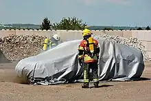 Ein brennendes Auto ist abgedeckt mit einer Brandbegrenzungsdecke, wenig Rauch ist zu sehen, zwei Feuerwehrleute mit Atemschutz beobachten die Situation.
