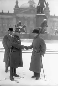 Schwarzweißes Seitenprofil von zwei lächelnden Männern, die sich die Hand geben. Der linke Mann trägt eine Militäruniform und der rechte Mann einen Melonenhut und einen Mantel. Er stützt sich auf einem Gehstock ab. Hinten links steht ein Mann, der ihnen zuschaut. Im Hintergrund sind ein Gebäude mit Kuppeldach und ein Denkmal mit Figuren.