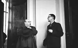 Dieses Bild zeigt auf der linken Seite Hanns Eisler, rechts Bertolt Brecht im Gespräch. Wie so oft, Brecht hält in seiner linken Hand eine Zigarre, Eisler hat einen Mantel an und steht mit verschränkten Armen. Das Bild selbst ist undatiert, muss aber nach 1950 aufgenommen worden sein und stammt aus dem deutschen Bundesarchiv.