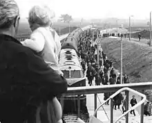 Mann mit Kleinkind auf dem Arm steht auf einer Brücke, die Kamera sieht an ihm vorbei auf einen Zug an einem Bahnsteig, dort steigen viele Arbeiter aus.