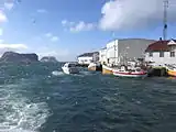Foto mit einigen Hafengebäuden auf der rechten Seite, einige Boote im Wasser davon und zwei Inseln im Hintergrund