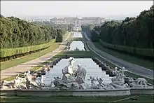 Frontale Farbfotografie einer rechteckigen Wasserfläche, die bis zum Hintergrund führt und an einem Schloss endet. Das Wasser wird durch zwei kleine Rasenflächen durchbrochen. An beiden Seiten sind Wege, Heckenreihen und Wälder. Im Vordergrund sind mehrere Skulpturen, die eine Szene darstellen.