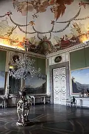 Farbige Eckansicht eines Innenraums mit einer Deckenmalerei, die Szenen aus dem Himmel und der Erde durch Putten und Blumenkränze verbindet. Von der Decke hängt ein Kronleuchter und unter ihm steht ein Ständer mit Schale. An den Wänden befinden sich drei Landschaftsgemälde, unter denen kleine Marmortische stehen. Über dem linken Spiegel und der rechten Tür hängen kleine, runde Gemälde.