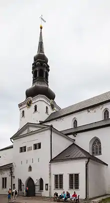 Frontale Farbfotografie von einer weißen Kirche mit schwarzer Dachhaube. Der Rundbogeneingang befindet sich in einem Vorbau mit Giebelfeld. Die Fassade ist schlicht gehalten. Im Vordergrund sitzen Menschen auf einer Bank.