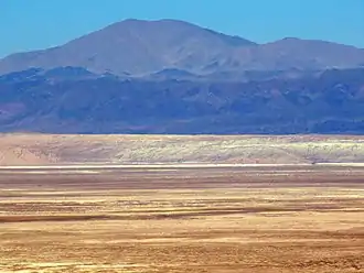 Die Cordillera Domeyko von den Hängen der Anden-Westkordillere aus gesehen. Auf 2.900 m ü. M. Höhe in 50 bis 70 km Entfernung in Richtung Westen betrachtet, erhebt sich hinter der Salar-de-Atacama-Senke (2.300 m ü. M.) und der Cordillera de la Sal der dunkle Steifen des El Bordo-Kliffs auf um die 3.000 m ü. M., vor dem höchsten Berg dem Cerro Quimal.