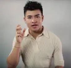 Porträtfoto von Chella Man vor weißem Hintergrund. Er hält die Finger der rechten Hand gespreizt und trägt ein weißes Hemd mit braunen Kreisen (bestehend aus je sechs Punkten), einen silbernen Ring am rechten Ringfinger sowie zwei schwarze ringförmige Tätowierungen am rechten Arm. Seine schwarzen Haare trägt er als bis zu den Ohren reichende Kurzhaarfrisur, wobei einzelne Haare nach oben abstehen.