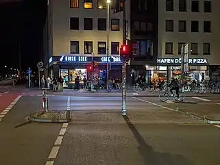 Junge Menschen cornern an einer Straßenecke im Hansaviertel in Münster. An der Straße befinden sich auch ein Kiosk und eine Imbissstube.