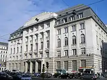 Seitliche Farbfotografie eines fünfgeschossigen Gebäudes mit einem ausgebauten Dachstuhl. Über dem Säuleneingang steht „Bank Austria Creditanstalt“. Der Mittelteil ist mit rechteckigen Säulen durchzogen und hat unter dem vierten Geschoss Figuren, die die Fenster stützen. Über den äußeren Fenstern befinden sich Kopfreliefs. Das ganze dritte Geschoss besteht aus Rundbogenfenstern. Auf dem Dach ist ein Staffelgiebel mit einem schwarzen Logo.