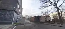 Eine fünf Meter breite Einfahrt mit geöffnetem Tor, links ein bedrohlich wirkendes Hochhaus, rechts ein Pförtnergebäude und ein blattloser großer Baum