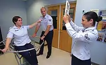 Eine Versuchsperson sitzt in einem Drehstuhl. Eine zweite Person hat gerade die Rotation des Stuhls gestoppt. Die orientierungslose Versuchsperson zeigt auf ein Zeichen, welches von einer dritten Person gehalten wird.