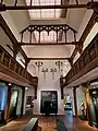 Blick in die Museumshalle des Dithmarscher Landesmuseums, an der Stirnseite zu sehen ist eine Medienstation zum Dithmarscher Landrecht, darauf zulaufend zwei graue Sitzmöbel, an den Seiten graue Ausstellungsmöbel und eine Etage höher die Ballustrade der Galerie