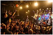 Farbfotografie von einem Konzert auf einer kleinen Bühne. Links jubelt das Publikum mit erhobenen Armen und rechts spielen vier Musiker, bestehend aus zwei Gitarristen, einem Schlagzeuger und einem Sänger. Der Sänger hält den Mikrofonständer ins Publikum. Im Hintergrund sind ein Graffiti-Bild, die Lautsprecher und Scheinwerfer.