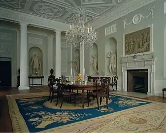 Farbige Eckansicht eines Innenraums mit einem runden Tisch und Stühlen, die in der Mitte auf einem blau-beige gemusterten Teppich stehen. Rechts befindet sich ein Kamin mit einem grauen Gemälde in weißem Rahmen darüber, das eine antike Szene darstellt. An beiden Wänden stehen in Nischen vier Marmorstatuen im griechischen Stil. Vor der linken Wand befinden sich Säulen und auf beiden Wänden sowie an der Decke sind weiße Ornamente angebracht.