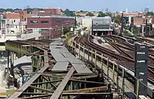 Viel Stahl und Stadt im Blickfeld: In der linken Bildhälfte das teils rostige, teils mit grünem Schutzanstrich versehene Tragwerk einer ehemaligen Hochbahnstrecke. Es basiert auf einem im Querschnitt rechteckigen Körper aus vier Trägern, die durch viele diagonale Verstrebungen gegenseitig zu einem stabilen Gesamttragwerk verbunden sind. Auf ihm lagen direkt die Schwellen und darauf die Schienen der Hochbahnstrecke. Man sieht hier ein solches Tragwerk nach links in der Bildmitte verlaufen und dort mechanisch abgeschnitten enden. Die rechts Bildhälfte wird von einem Rampenbauwerk mit vier Gleisen bestimmt, entlang dem die Betrachterin abwärts auf die nebeneinander angeordneten Tunnelportale der Culver Line blickt. An den Seiten der Rampe sind Straßen, Laternen, Schilder und Gebäude zu sehen, in der Ferne einzelne Hochhäuser vor diesigem Himmel.