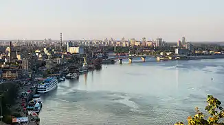 Von der rechten unteren Fotoecke bis zur Mitte erstreckt sich der Fluss. Daran schmiegt sich die Stadt halbkreisförmig von unten links nach rechts. Einige Schiffe und Fabriken sind links zu erkennen, in der Mitte befindet sich eine Autobrücke und in der Ferne sind Wolkenkratzer.