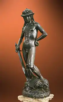 Frontale Farbfotografie einer nackten Bronzefigur, die nur einen Hut und Stiefel trägt. Seine rechte Hand hält ein nach unten gerichtetes Schwert und seine linke Hand ist an der Hüfte angewinkelt. Er steht auf Trümmern, die sich wiederum auf einem hellen Sockel befinden.
