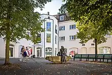 Fotos eines von Häusern gesäumten Platzes mit einer halbrunden Bankreihe um ein Denkmal einer Bronzefigur, die auf einem Stein sitzt