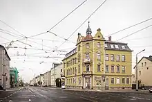 Foto einer Kreuzung mit Trambahnschienen und Oberleitungen aus bzw. in alle Fahrtrichtungen. Im Hintergrund ein ockerfarbenes Haus.