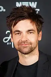 Drew Tarver steht vor einer schwarzen Wand mit weißen, verdeckten Schriftzügen. Er trägt ein schwarzes T-Shirt, ein schwarzes Jackett und einen kurzen Stoppelbart. Seine braunen Haare trägt er als bis zu den Ohren reichende, verwuschelte Kurzhaarfrisur mit einigen hochgestellten Haarspitzen.