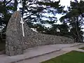 Das Foto eines Denkmals, das vor Bäumen steht. Das Denkmal ist eine Bruchsteinmauer, die einen leichten Bogen beschreibt und auf der linken Seite höher ist als auf der rechten. An der Stelle, an der die Mauer am höchsten ist, befindet sich ein weißes Kreuz auf der Mauer. Am rechten Ende der Mauer ist eine Plakette mit einer Beschriftung zu erkennen. Diese ist allerdings nicht zu lesen. Vor dem Denkmal befindet sich eine Wiese.